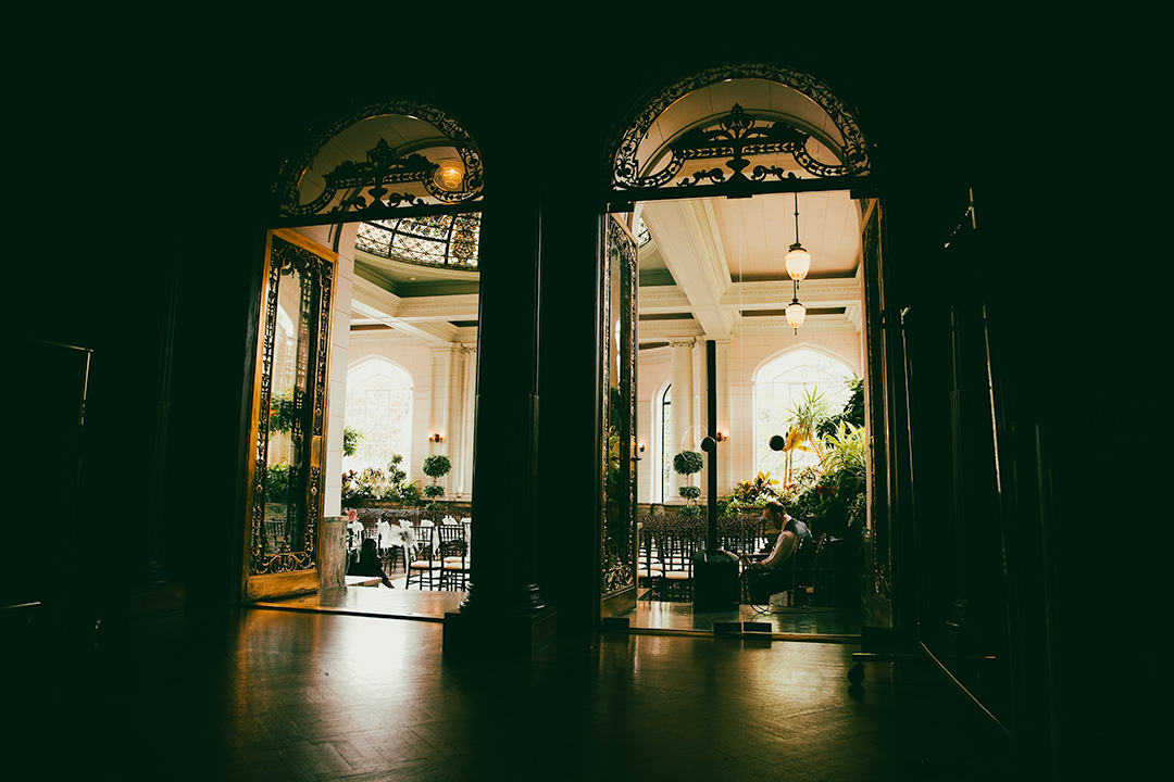 View looking through two large ornate doors into a room with many chairs. A musician is seated on the right.  